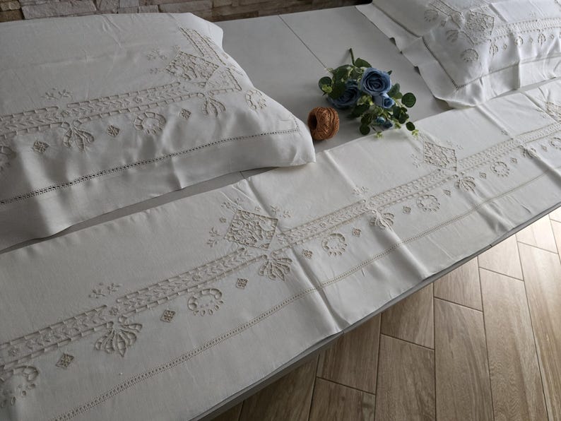 May include: White embroidered bedding set with a pillowcase, runner, and another pillowcase. The set features detailed floral and geometric patterns. A blue floral arrangement and a decorative wooden ball are placed on the bedding.