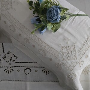 May include: White embroidered pillowcases with detailed floral and geometric patterns. A bouquet of blue flowers sits on the pillow. The pillowcases have cutwork and decorative stitching, giving a vintage look.