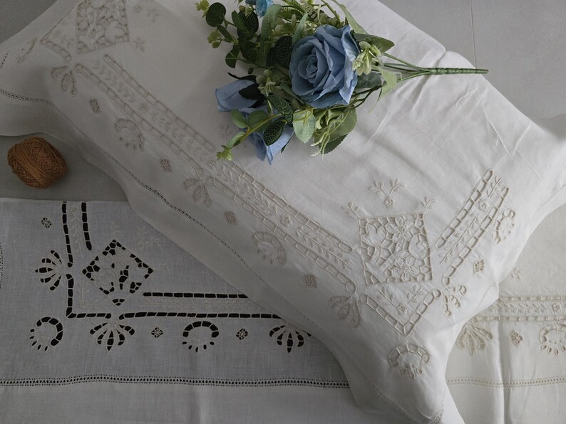 May include: White embroidered pillowcases with detailed floral and geometric patterns. A bouquet of blue flowers sits on the pillow. The pillowcases have cutwork and decorative stitching, giving a vintage look.