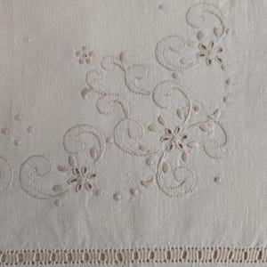 K&ouml;nnte beinhalten: Cremefarbene Leinentischdecke mit gestickten Blumen- und Wirbelmustern in hellem Beige. Der Rand ist mit einer dekorativen Bord&uuml;re versehen. Das Design erinnert an einen Vintage-Stil.