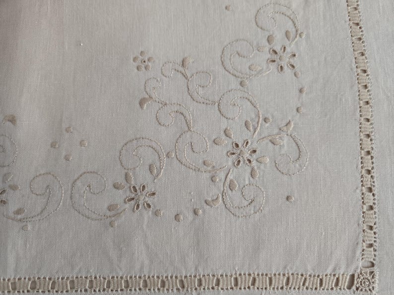 K&ouml;nnte beinhalten: Cremefarbene Leinentischdecke mit gestickten Blumen- und Wirbelmustern in hellem Beige. Der Rand ist mit einer dekorativen Bord&uuml;re versehen. Das Design erinnert an einen Vintage-Stil.