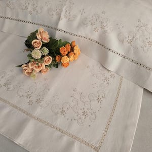K&ouml;nnte beinhalten: Wei&szlig;es besticktes Bettw&auml;sche-Set mit floralen und Rankenmustern. Das Set enth&auml;lt einen Kissenbezug und ein Laken mit dekorativem Rand. Ein Strau&szlig; aus Pfirsich- und Orangenrosen liegt auf dem Laken.
