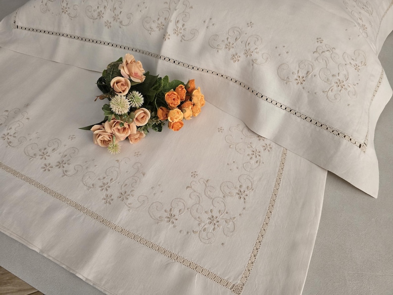K&ouml;nnte beinhalten: Wei&szlig;es besticktes Bettw&auml;sche-Set mit floralen und Rankenmustern. Das Set enth&auml;lt einen Kissenbezug und ein Laken mit dekorativem Rand. Ein Strau&szlig; aus Pfirsich- und Orangenrosen liegt auf dem Laken.