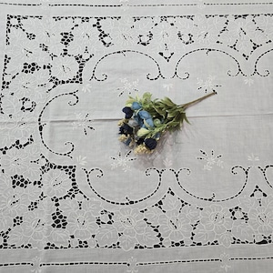 May include: A white, rectangular tablecloth with intricate floral and leaf cutwork designs. The edges are finished with a delicate, stitched border. A small bouquet of blue and white flowers rests on the cloth.