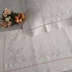 K&ouml;nnte beinhalten: Cremefarbenes besticktes Leinen-Bettw&auml;sche-Set. Das Set beinhaltet einen Kissenbezug und ein Laken, beide mit zarten floralen und scrollartigen Stickereien an den R&auml;ndern. Ein kleiner Strau&szlig; Pfirsich- und Orangenbl&uuml;ten liegt auf dem Bett.