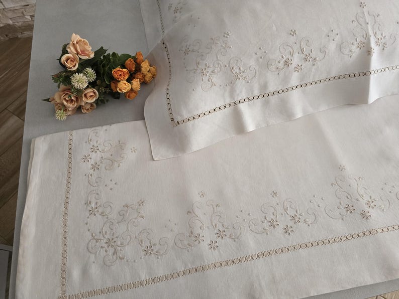 K&ouml;nnte beinhalten: Cremefarbenes besticktes Leinen-Bettw&auml;sche-Set. Das Set beinhaltet einen Kissenbezug und ein Laken, beide mit zarten floralen und scrollartigen Stickereien an den R&auml;ndern. Ein kleiner Strau&szlig; Pfirsich- und Orangenbl&uuml;ten liegt auf dem Bett.