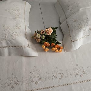 K&ouml;nnte beinhalten: Cremefarbenes besticktes Bettw&auml;sche-Set mit zwei Kissenbez&uuml;gen und einem Spannbetttuch. Die Kissenbez&uuml;ge und das Laken weisen zarte Blumen- und Rankenstickereien auf. Ein Strau&szlig; aus orangefarbenen und rosa Rosen liegt auf dem Bett.