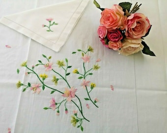 May include: White linen tablecloth with delicate pink and green embroidered flowers. A bouquet of pink roses is placed on the tablecloth.