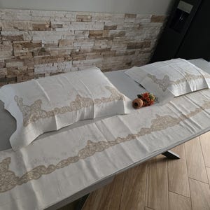 May include: Cream-colored bedding set featuring two pillowcases and a long runner, all adorned with a delicate, ornate beige lace design. The set is displayed on a gray surface, with a neutral-toned stone wall in the background.