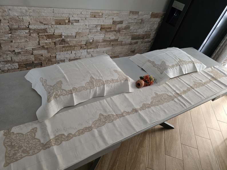May include: Cream-colored bedding set featuring two pillowcases and a long runner, all adorned with a delicate, ornate beige lace design. The set is displayed on a gray surface, with a neutral-toned stone wall in the background.
