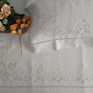 K&ouml;nnte beinhalten: Wei&szlig;es besticktes Bettw&auml;sche-Set mit floralen und scrollartigen Designs. Das Set beinhaltet einen Kissenbezug und ein Laken, beide mit zarter Stickerei und einem dekorativen Rand. Ein Strau&szlig; aus orangefarbenen und pfirsichfarbenen Rosen liegt auf dem Laken und f&uuml;gt einen Farbtupfer hinzu.