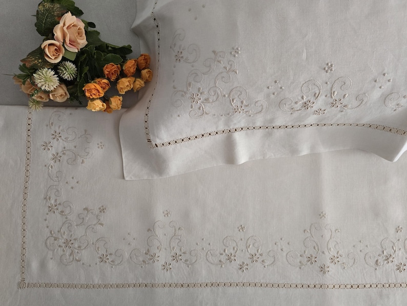 K&ouml;nnte beinhalten: Wei&szlig;es besticktes Bettw&auml;sche-Set mit floralen und scrollartigen Designs. Das Set beinhaltet einen Kissenbezug und ein Laken, beide mit zarter Stickerei und einem dekorativen Rand. Ein Strau&szlig; aus orangefarbenen und pfirsichfarbenen Rosen liegt auf dem Laken und f&uuml;gt einen Farbtupfer hinzu.