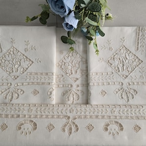 May include: Cream-coloured embroidered linens with floral and geometric designs. The linens have detailed cutwork and decorative borders. A bouquet of blue flowers sits above the folded textiles, adding a touch of colour and elegance to the display.