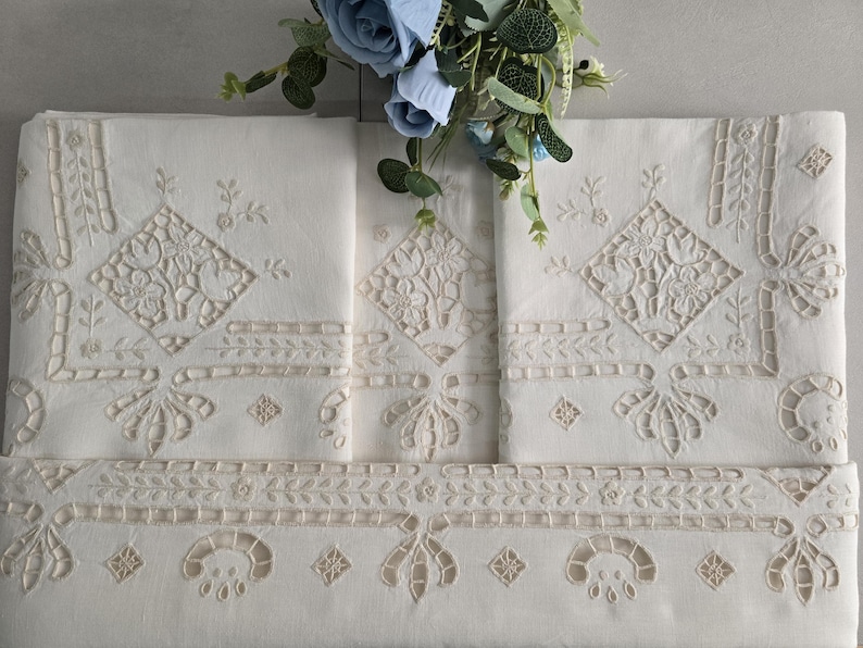 May include: Cream-coloured embroidered linens with floral and geometric designs. The linens have detailed cutwork and decorative borders. A bouquet of blue flowers sits above the folded textiles, adding a touch of colour and elegance to the display.