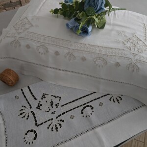 May include: White embroidered pillowcases and a matching runner with a floral design. The pillowcases have a detailed, symmetrical pattern in a lighter shade of white. A small, woven brown ball sits near the pillowcase. Blue flowers are on top.