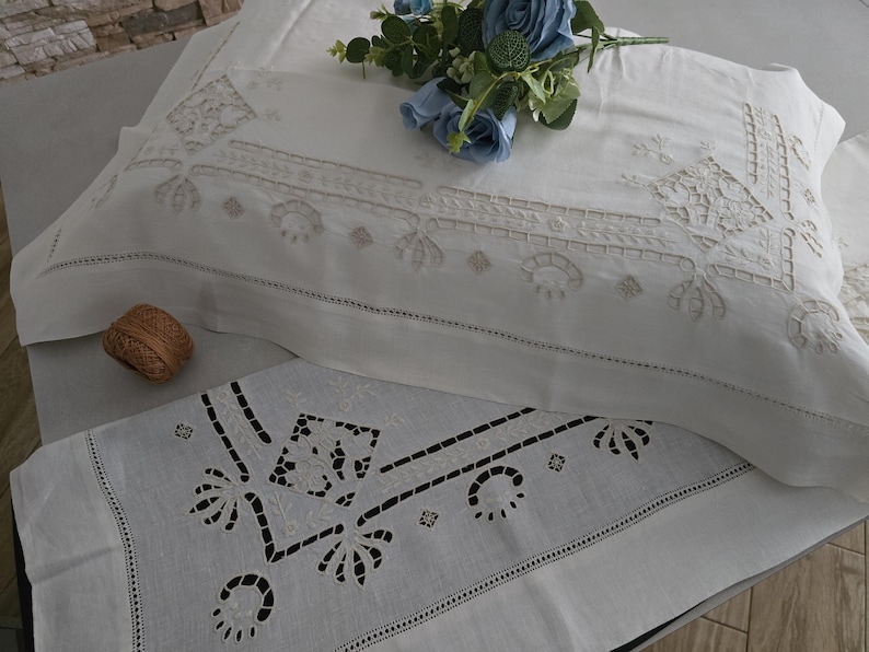 May include: White embroidered pillowcases and a matching runner with a floral design. The pillowcases have a detailed, symmetrical pattern in a lighter shade of white. A small, woven brown ball sits near the pillowcase. Blue flowers are on top.