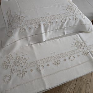 May include: Cream-coloured embroidered pillowcase and runner set. The set features detailed floral and geometric patterns in a lighter cream colour. The pillowcase and runner are made of a soft, likely linen or cotton fabric, and are suitable for a bedroom.