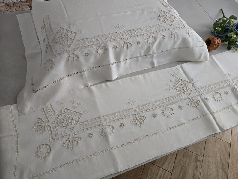 May include: Cream-coloured embroidered pillowcase and runner set. The set features detailed floral and geometric patterns in a lighter cream colour. The pillowcase and runner are made of a soft, likely linen or cotton fabric, and are suitable for a bedroom.