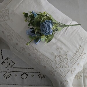 May include: White linen pillowcases with embroidered floral and geometric designs. A bouquet of blue roses and greenery rests on the pillow. A decorative linen runner with cut-out patterns is also visible.