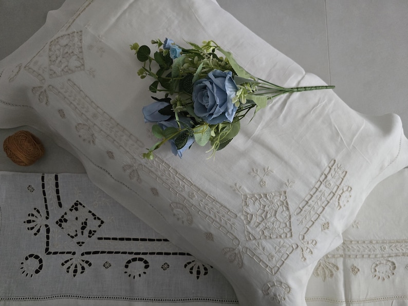 May include: White linen pillowcases with embroidered floral and geometric designs. A bouquet of blue roses and greenery rests on the pillow. A decorative linen runner with cut-out patterns is also visible.