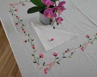 May include: White tablecloth with pink embroidered flowers and green leaves. A white napkin is folded on the tablecloth. A potted orchid with pink flowers is in the background.