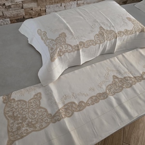 May include: Cream-colored bedding set featuring a pillowcase and a sheet, both adorned with intricate beige lace detailing along the edges. The set is displayed on a light gray surface, showcasing the delicate embroidery and texture.
