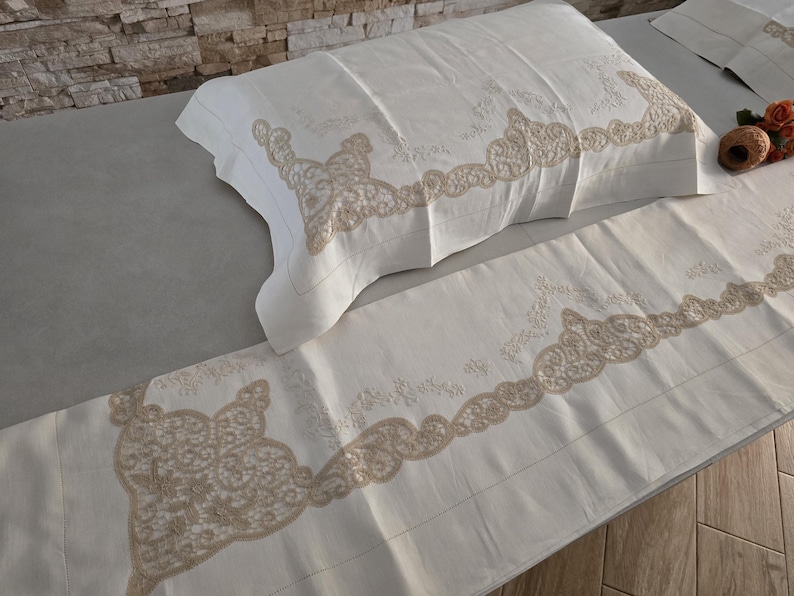 May include: Cream-colored bedding set featuring a pillowcase and a sheet, both adorned with intricate beige lace detailing along the edges. The set is displayed on a light gray surface, showcasing the delicate embroidery and texture.