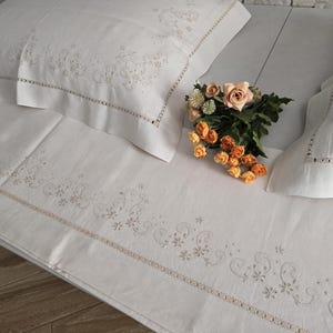 K&ouml;nnte beinhalten: Wei&szlig;es besticktes Bettw&auml;sche-Set mit floralen und Ranken-Details. Das Set beinhaltet ein Spannbetttuch, zwei Kissenbez&uuml;ge und ein Bettlaken. Ein Strau&szlig; aus orangefarbenen und pfirsichfarbenen Rosen liegt auf dem Bett.