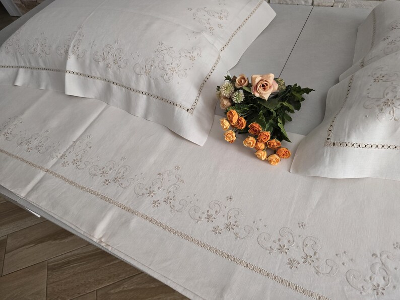 K&ouml;nnte beinhalten: Wei&szlig;es besticktes Bettw&auml;sche-Set mit floralen und Ranken-Details. Das Set beinhaltet ein Spannbetttuch, zwei Kissenbez&uuml;ge und ein Bettlaken. Ein Strau&szlig; aus orangefarbenen und pfirsichfarbenen Rosen liegt auf dem Bett.