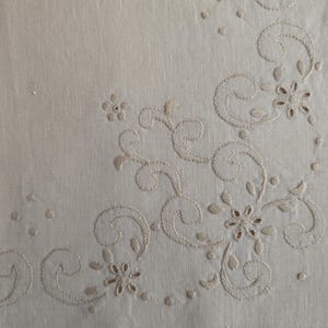 K&ouml;nnte beinhalten: Cremefarbener Leinenstoff mit gesticktem Blumen- und Rankenmuster. Die Stickerei zeigt zarte Blumen, Bl&auml;tter und wirbelnde Muster in passendem cremefarbenem Faden. Der Stoff hat eine subtile Textur.