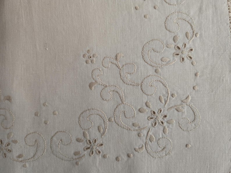 K&ouml;nnte beinhalten: Cremefarbener Leinenstoff mit gesticktem Blumen- und Rankenmuster. Die Stickerei zeigt zarte Blumen, Bl&auml;tter und wirbelnde Muster in passendem cremefarbenem Faden. Der Stoff hat eine subtile Textur.