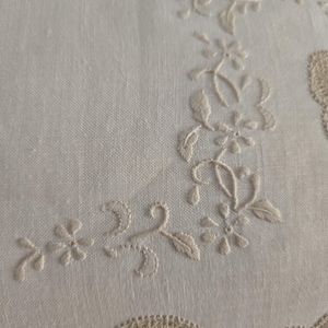 May include: Close-up of a white linen textile with delicate floral embroidery. The embroidery features a repeating pattern of small flowers and leaves in a light beige color, creating a subtle, elegant design. The fabric has a fine weave.
