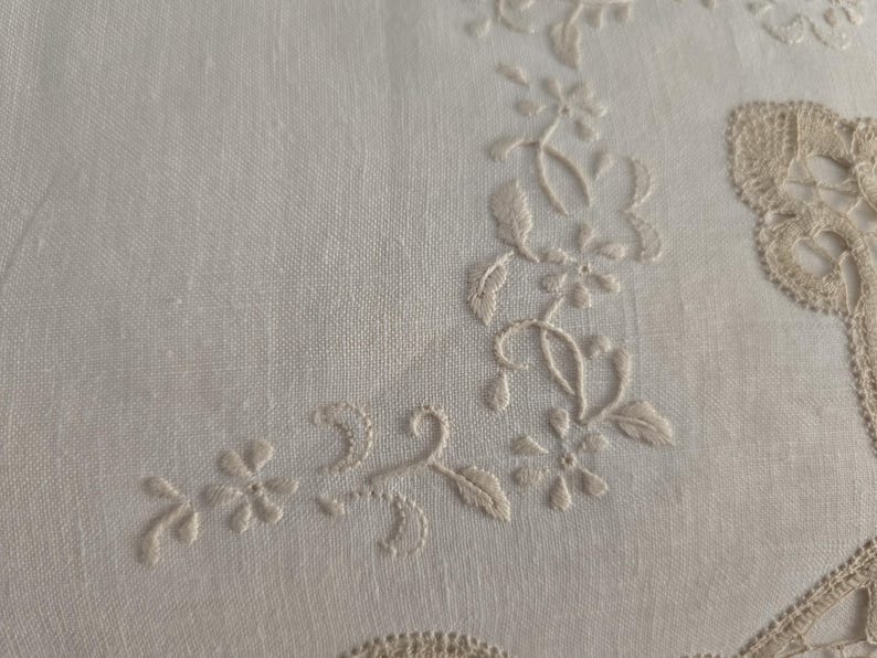 May include: Close-up of a white linen textile with delicate floral embroidery. The embroidery features a repeating pattern of small flowers and leaves in a light beige color, creating a subtle, elegant design. The fabric has a fine weave.