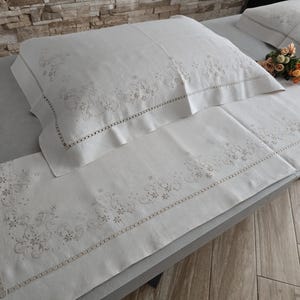 K&ouml;nnte beinhalten: Wei&szlig;es Leinen-Bettw&auml;sche-Set mit gestickten Blumenmustern. Das Set enth&auml;lt einen Kissenbezug, einen Bettl&auml;ufer und einen zweiten Kissenbezug. Die Stickerei ist in einer zarten, hellen Farbe gehalten und das Set ist ordentlich auf einem Bett angeordnet.