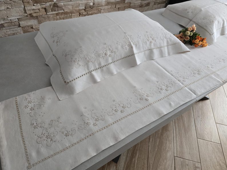K&ouml;nnte beinhalten: Wei&szlig;es Leinen-Bettw&auml;sche-Set mit gestickten Blumenmustern. Das Set enth&auml;lt einen Kissenbezug, einen Bettl&auml;ufer und einen zweiten Kissenbezug. Die Stickerei ist in einer zarten, hellen Farbe gehalten und das Set ist ordentlich auf einem Bett angeordnet.