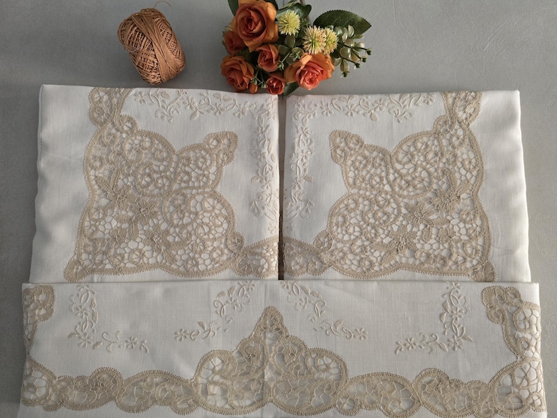 May include: White linen bedding set with intricate beige lace detailing. The set includes pillowcases and a larger piece, possibly a duvet cover or sheet, all adorned with floral embroidery and lace. A ball of brown thread and orange flowers are also present.