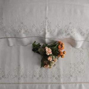 K&ouml;nnte beinhalten: Wei&szlig;es Leinen-Bettw&auml;sche-Set mit gestickten floralen Details und einem dekorativen Rand. Ein Strau&szlig; aus pfirsichfarbenen und orangefarbenen Blumen liegt auf der Bettw&auml;sche. Kissenbezug und Laken weisen zarte N&auml;hte auf.