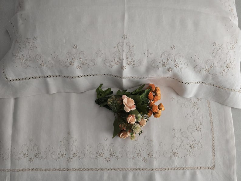 K&ouml;nnte beinhalten: Wei&szlig;es Leinen-Bettw&auml;sche-Set mit gestickten floralen Details und einem dekorativen Rand. Ein Strau&szlig; aus pfirsichfarbenen und orangefarbenen Blumen liegt auf der Bettw&auml;sche. Kissenbezug und Laken weisen zarte N&auml;hte auf.