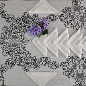 May include: A white tablecloth with intricate black lace detailing and matching white napkins. The lace design features floral patterns and decorative borders. A small bouquet of purple flowers adds a touch of color to the scene.