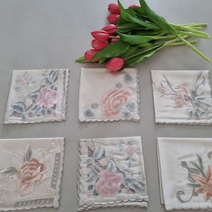 May include: Six white embroidered handkerchiefs with floral designs. The handkerchiefs have a scalloped edge and are arranged in two rows of three. The handkerchiefs are on a light grey surface.