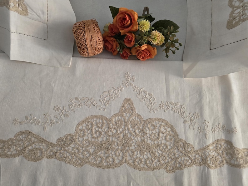 May include: A white textile with intricate beige lace and floral embroidery. A ball of tan thread and a bouquet of orange roses and yellow flowers are placed on the textile. The overall theme is vintage textiles.