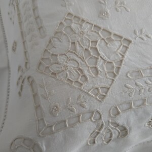 May include: Close-up of a white, embroidered textile with intricate floral and geometric patterns. The design features openwork details and delicate stitching, creating a vintage or antique aesthetic. The fabric appears to be linen or a similar natural fibre.
