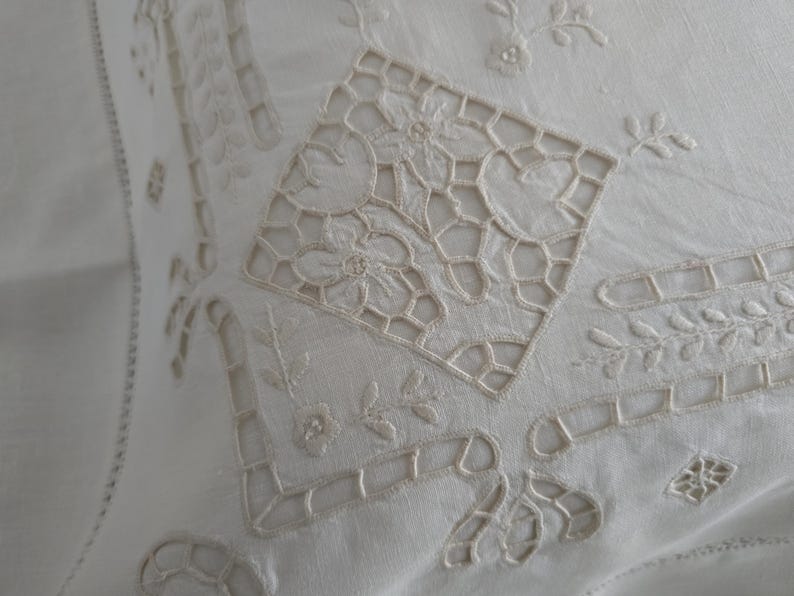 May include: Close-up of a white, embroidered textile with intricate floral and geometric patterns. The design features openwork details and delicate stitching, creating a vintage or antique aesthetic. The fabric appears to be linen or a similar natural fibre.