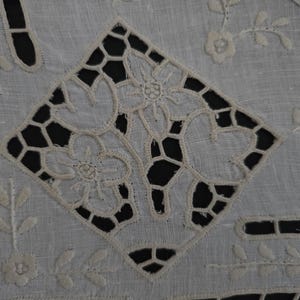 May include: Close-up of a white, embroidered textile featuring a central diamond pattern with openwork and floral designs. The textile has additional floral and leaf motifs around the edges, creating a delicate, detailed appearance.