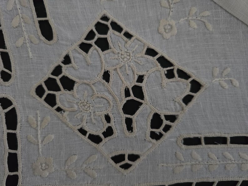 May include: Close-up of a white, embroidered textile featuring a central diamond pattern with openwork and floral designs. The textile has additional floral and leaf motifs around the edges, creating a delicate, detailed appearance.