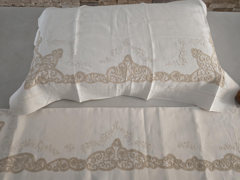 May include: White bedding set featuring a pillowcase and a sheet, both adorned with intricate beige lace detailing along the edges. The lace design adds a touch of elegance to the otherwise plain white fabric.