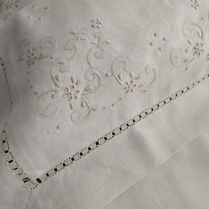 K&ouml;nnte beinhalten: Nahaufnahme eines wei&szlig;en Leinen-Kissenbezugs mit aufwendiger floraler Stickerei in hellem Beige. Der Kissenbezug hat einen zarten, durchbrochenen Rand, der einen Hauch von Eleganz verleiht. Das Design umfasst wirbelnde Muster und kleine Blumenmotive.