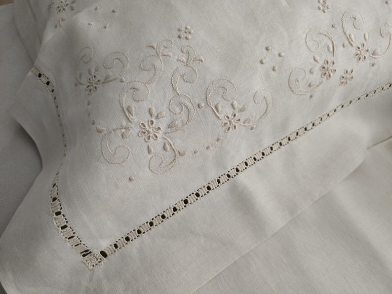 K&ouml;nnte beinhalten: Nahaufnahme eines wei&szlig;en Leinen-Kissenbezugs mit aufwendiger floraler Stickerei in hellem Beige. Der Kissenbezug hat einen zarten, durchbrochenen Rand, der einen Hauch von Eleganz verleiht. Das Design umfasst wirbelnde Muster und kleine Blumenmotive.