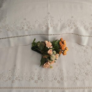 K&ouml;nnte beinhalten: Wei&szlig;es Leinen-Bettw&auml;sche-Set mit gestickten Blumen- und Rankenmustern. Das Set beinhaltet einen Kissenbezug und ein Laken mit dekorativen Bord&uuml;ren. Ein kleiner Strau&szlig; aus Pfirsich- und orangefarbenen Blumen liegt auf dem Laken.