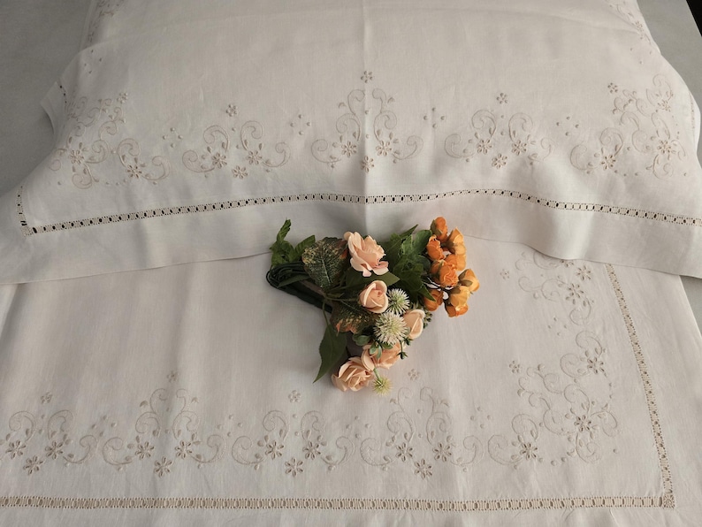 K&ouml;nnte beinhalten: Wei&szlig;es Leinen-Bettw&auml;sche-Set mit gestickten Blumen- und Rankenmustern. Das Set beinhaltet einen Kissenbezug und ein Laken mit dekorativen Bord&uuml;ren. Ein kleiner Strau&szlig; aus Pfirsich- und orangefarbenen Blumen liegt auf dem Laken.
